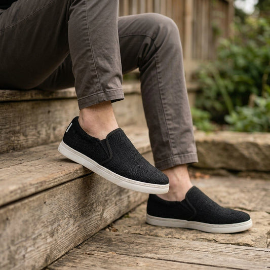 Person wearing black hemp vegan slip-on shoes with white rubber soles sitting on wooden steps.