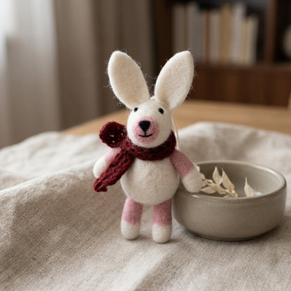 Handmade wool felt bunny ornaments in assorted scarf designs a red scarf on a textured surface with a bowl in the background