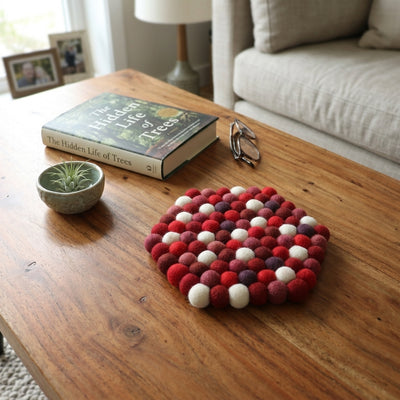 Decorative felt ball coaster on a wooden coffee table with a book and small plant.