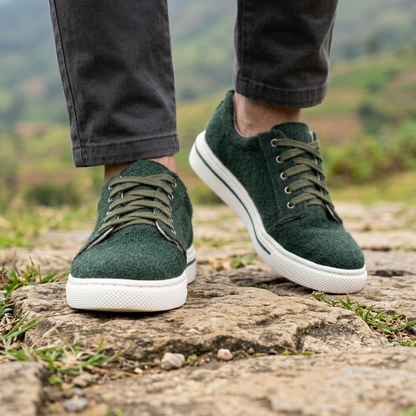 A person wearing dark green low-top sneakers with white soles, standing on a rocky outdoor path with blurred green hills in the background.