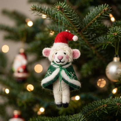 A handmade felt Winter Wanderer Sheep Christmas ornament wearing a red Santa hat and embroidered green snowflake cape, hanging from a pine branch with warm bokeh lights behind it.

